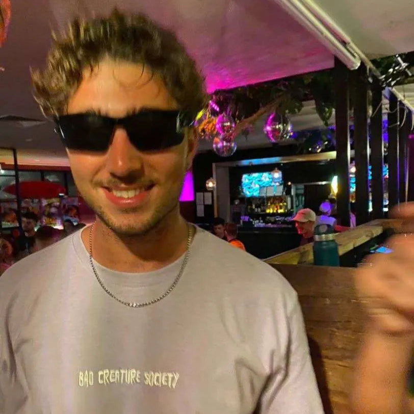 A Man with shades on wearing a Lilac T in a bar - he is smiling.
