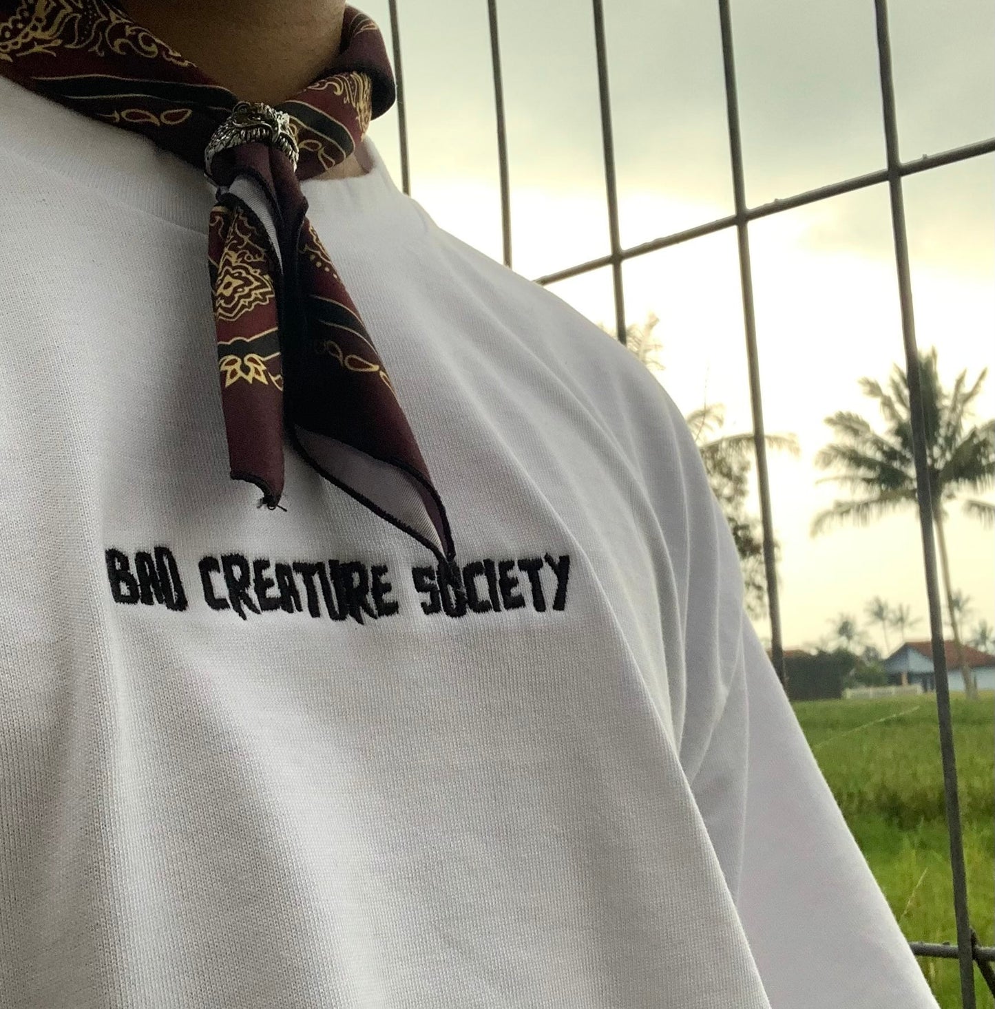 a close up of the white tee, with the wearer also wearing a patterned neck-scarf. Outside in front of a metal cage.