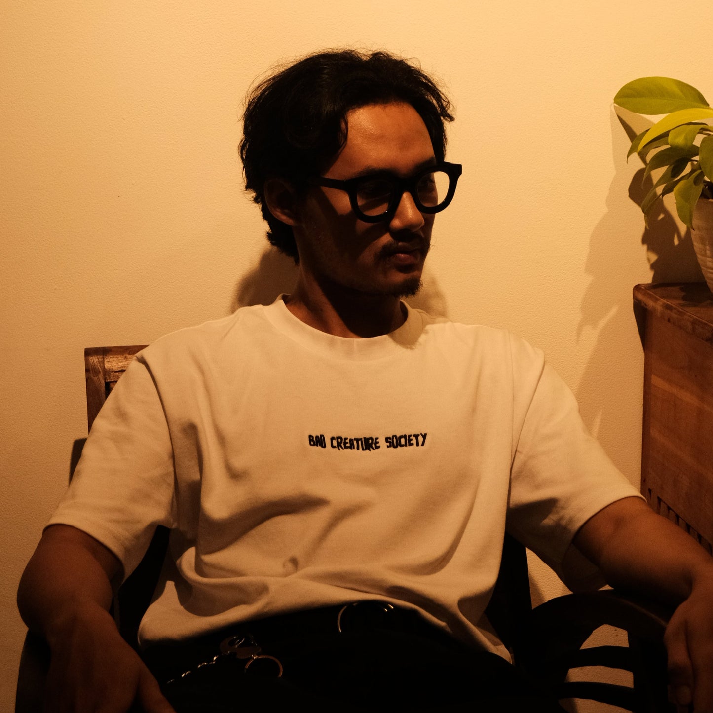 a person with glasses sitting down wearing a white tee with 'Bad Creature Society' embroidered on it, in a dimly lit room.