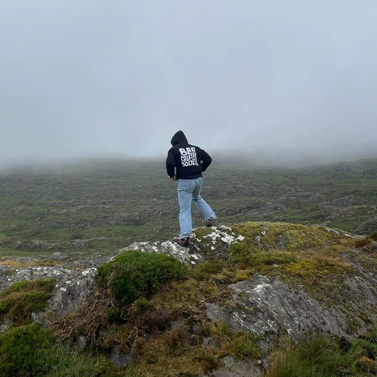Person wearing a hoodie with text standing on a rocky outcrop in a foggy landscape
