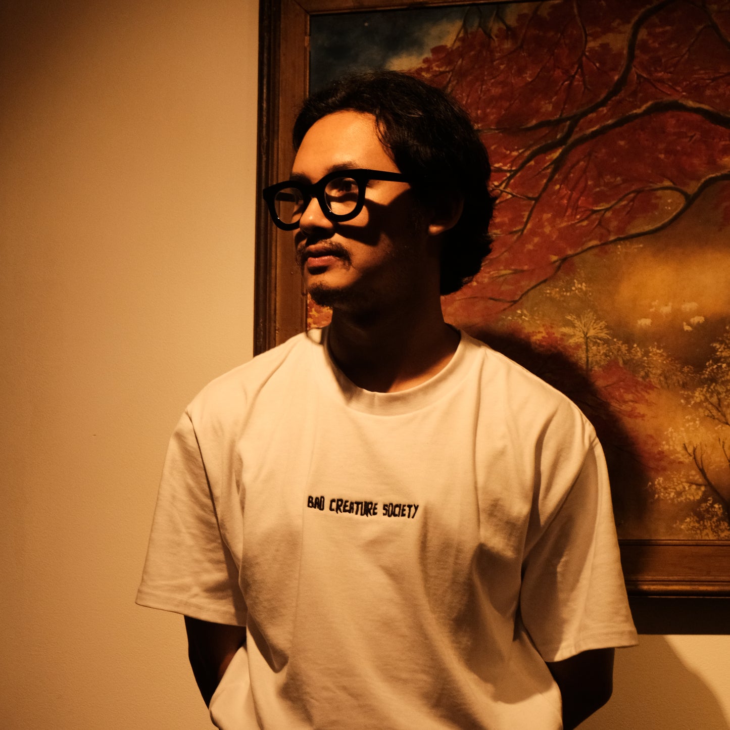 a person with glasses wearing a white tee with 'Bad Creature Society' embroidered on it, in a dimly lit room.