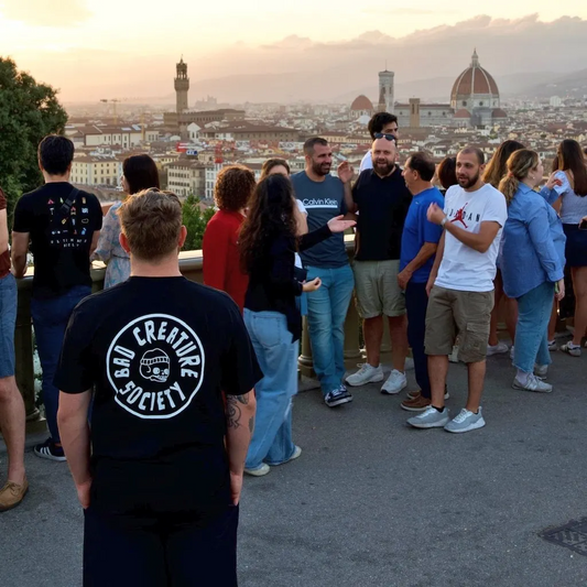 Group of people standing on a rooftop overlooking a cityscape with a sunset. At the forefront, the model is wearing the black Bad Creature Society 'Penny' Logo tee. 